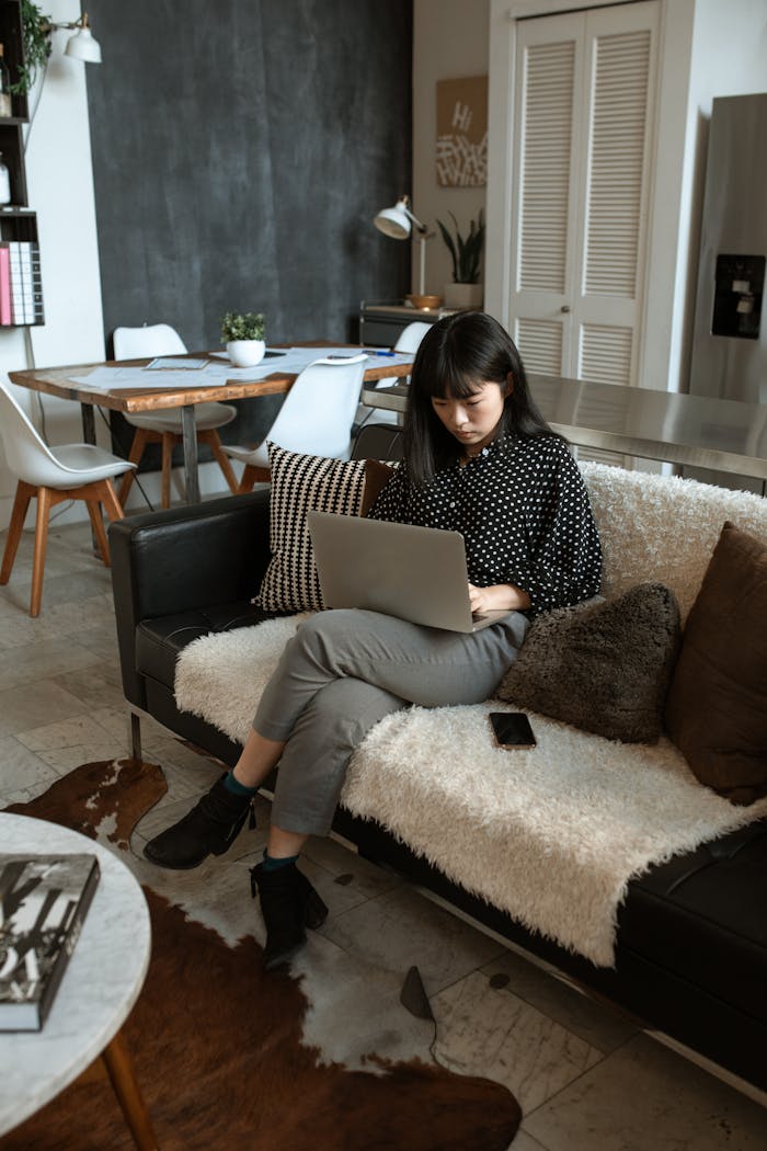 Asian woman sitting on a sofa, working on a laptop in a modern home interior setting.